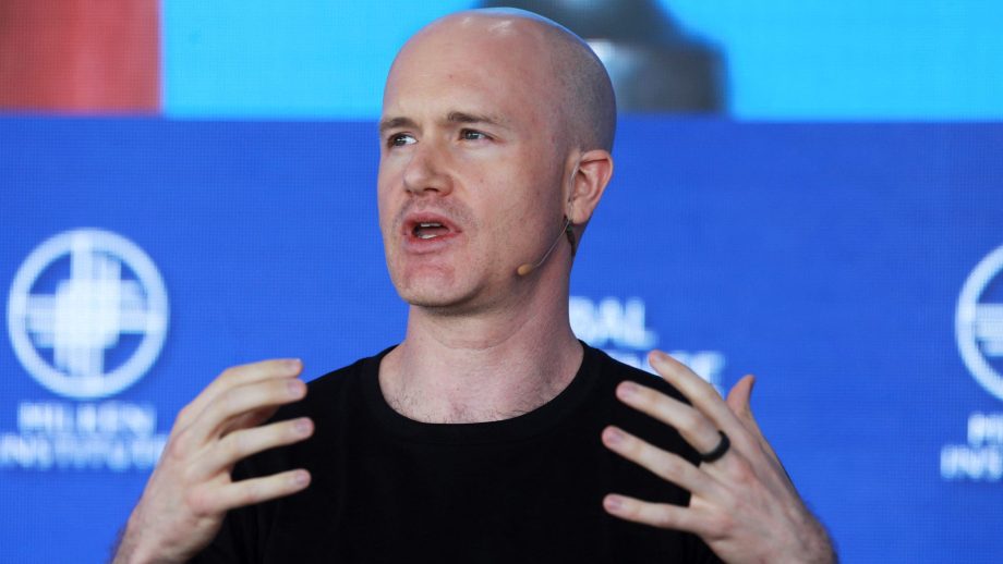 Ein kahlköpfiger Mann mit schwarzem Hemd und Headset-Mikrofon gestikuliert mit beiden Händen, während er auf der Bühne vor einem blauen Hintergrund mit Coinbase-Logos und weißem Text spricht.