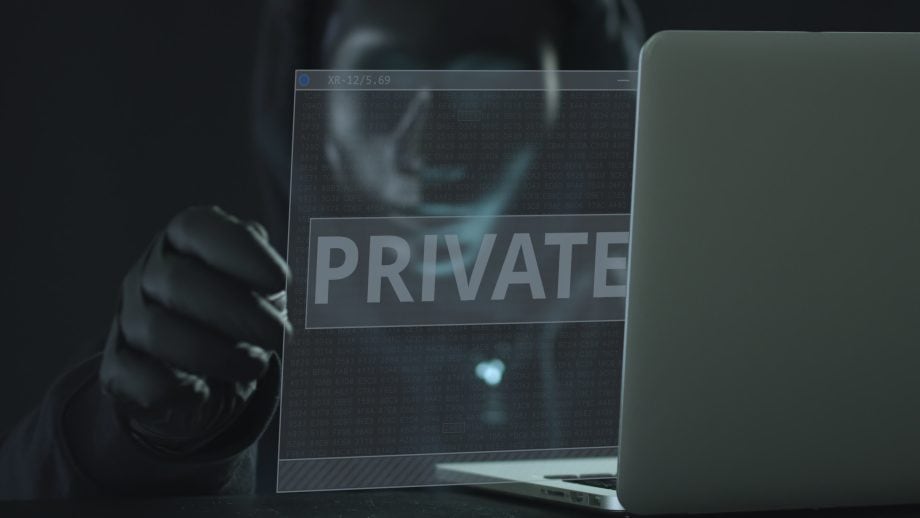 Eine Person in einem dunklen Kapuzenpullover sitzt hinter einem Laptop und hält einen transparenten Bildschirm in der Hand, auf dem in großen Buchstaben das Wort PRIVAT angezeigt wird, während auf dem Display schwach ein Bitcoin-Symbol leuchtet.
