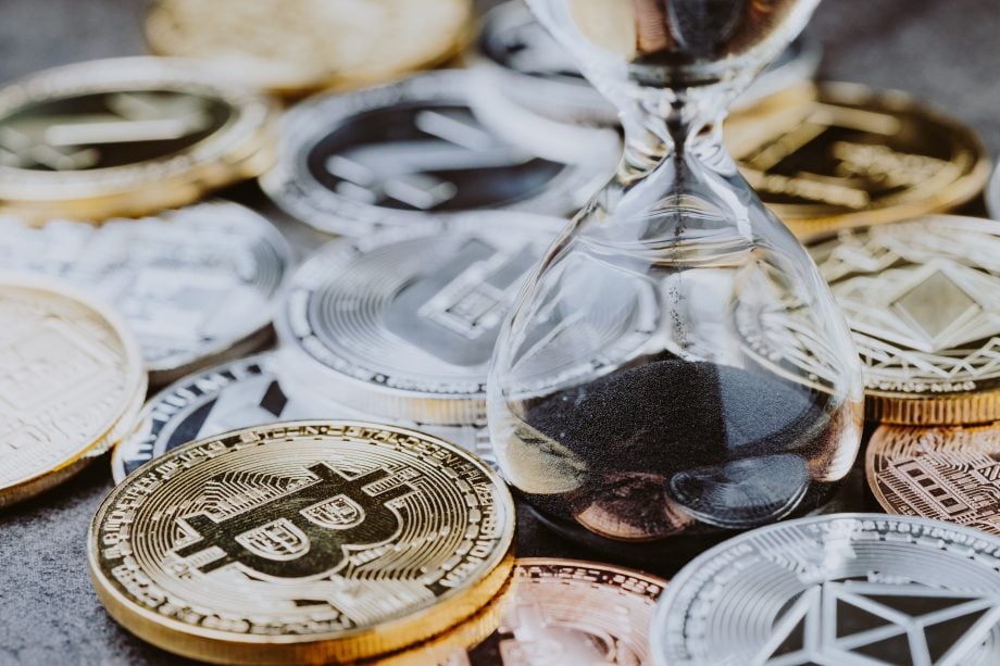 Eine Sanduhr steht auf einer Fläche, die mit verschiedenen Krypto-Münzen, darunter Bitcoin, Ethereum und Litecoin, bestückt ist.
