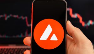 Eine Hand hält ein Smartphone mit einem roten Kreissymbol und zwei weißen Dreiecken, das dem AVAX-Symbol ähnelt. Im Hintergrund ist auf einem Laptop-Bildschirm ein unscharfes Kryptowährungsdiagramm zu sehen.