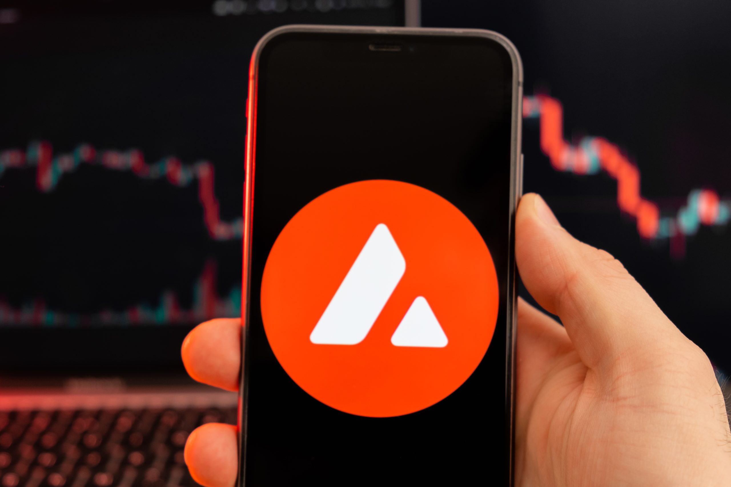 Eine Hand hält ein Smartphone mit einem roten Kreissymbol und zwei weißen Dreiecken, das dem AVAX-Symbol ähnelt. Im Hintergrund ist auf einem Laptop-Bildschirm ein unscharfes Kryptowährungsdiagramm zu sehen.