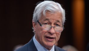 Ein älterer Mann mit grauem Haar und Brille, Anzug und Krawatte, spricht nachdenklich. Der Hintergrund ist unscharf und suggeriert ein professionelles Ambiente – möglicherweise eine Veranstaltung oder Konferenz von JPMorgan.