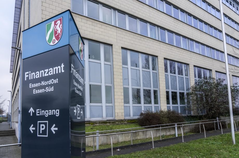 Ein Gebäude mit großen Fenstern und einem Schild mit der Aufschrift Finanzamt Essen-NordOst Essen-Süd, mit einer Wegbeschreibung zum Eingang, Parkmöglichkeiten und Informationen über Krypto-Steuerdienstleistungen.