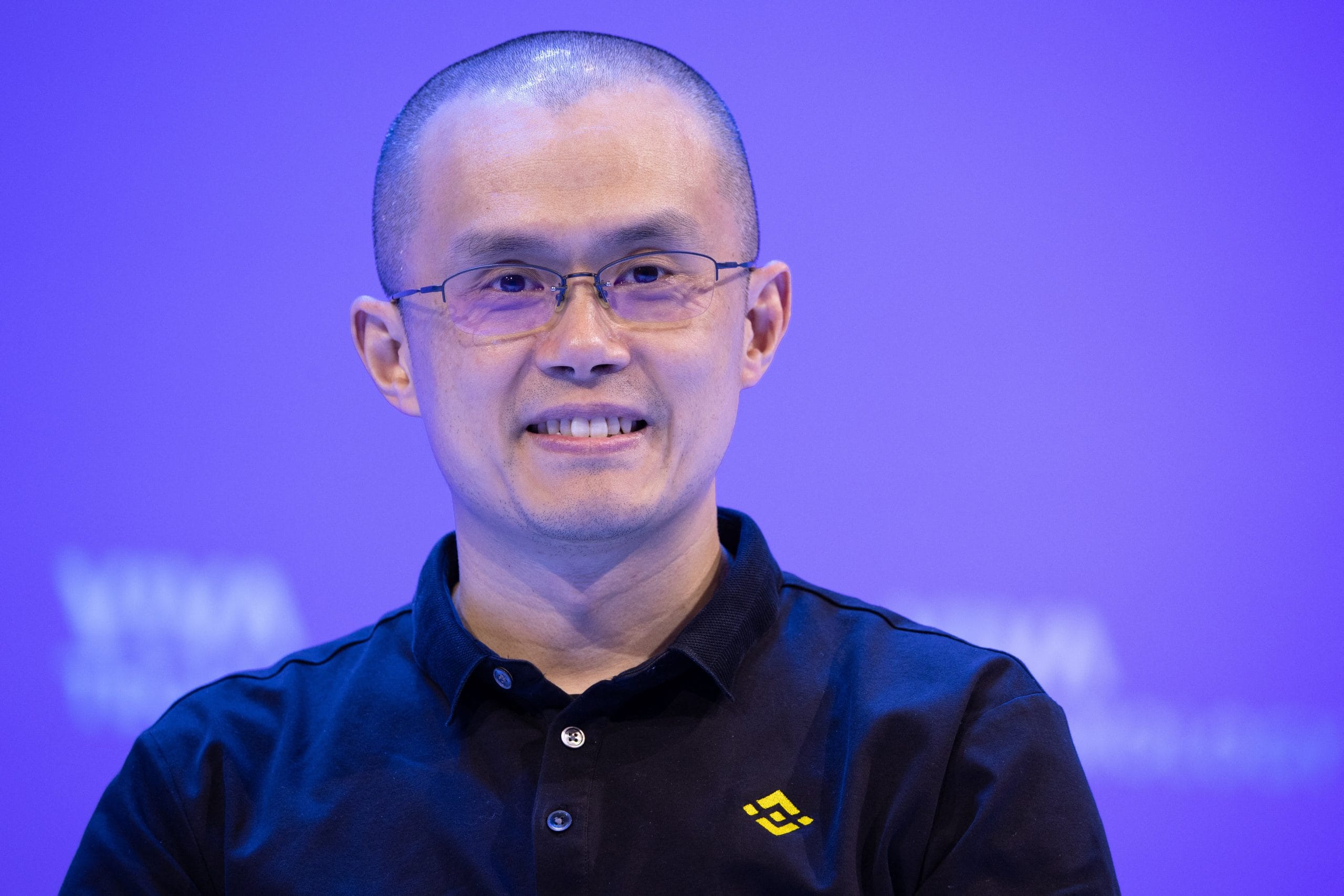 Eine Person mit Brille und schwarzem Hemd mit einem gelben Binance-Logo sitzt vor einem bläulich-violetten Hintergrund.