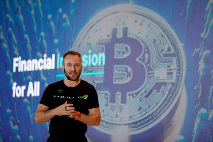 Eine Person in einem schwarzen Hemd steht vor einem großen Bildschirm, auf dem neben dem Tether-Logo das Bitcoin-Symbol angezeigt wird, was die Botschaft „Finanzielle Inklusion für alle“ unterstreicht.