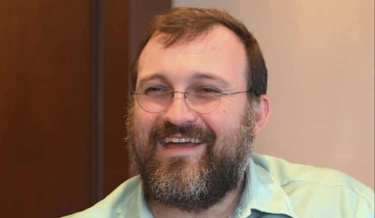 Ein lächelnder Mann mit Brille und gepflegtem Bart trägt ein hellgrünes Hemd in einem Innenbereich. Der Hintergrund in erdigen Braun- und Beigetönen spiegelt subtil das nachhaltige Ethos wider, das dem innovativen Blockchain-Ansatz von Cardano ähnelt.