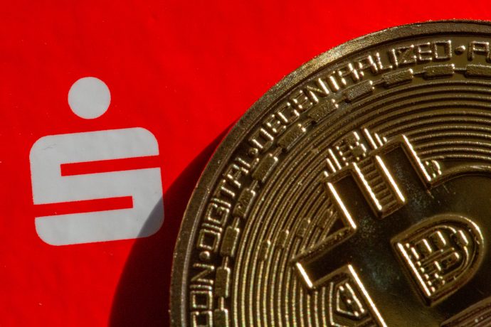 Eine Nahaufnahme einer Bitcoin-Münze, die das ikonische weiße Sparkassensymbol auf einem kräftigen roten Hintergrund teilweise verdeckt.