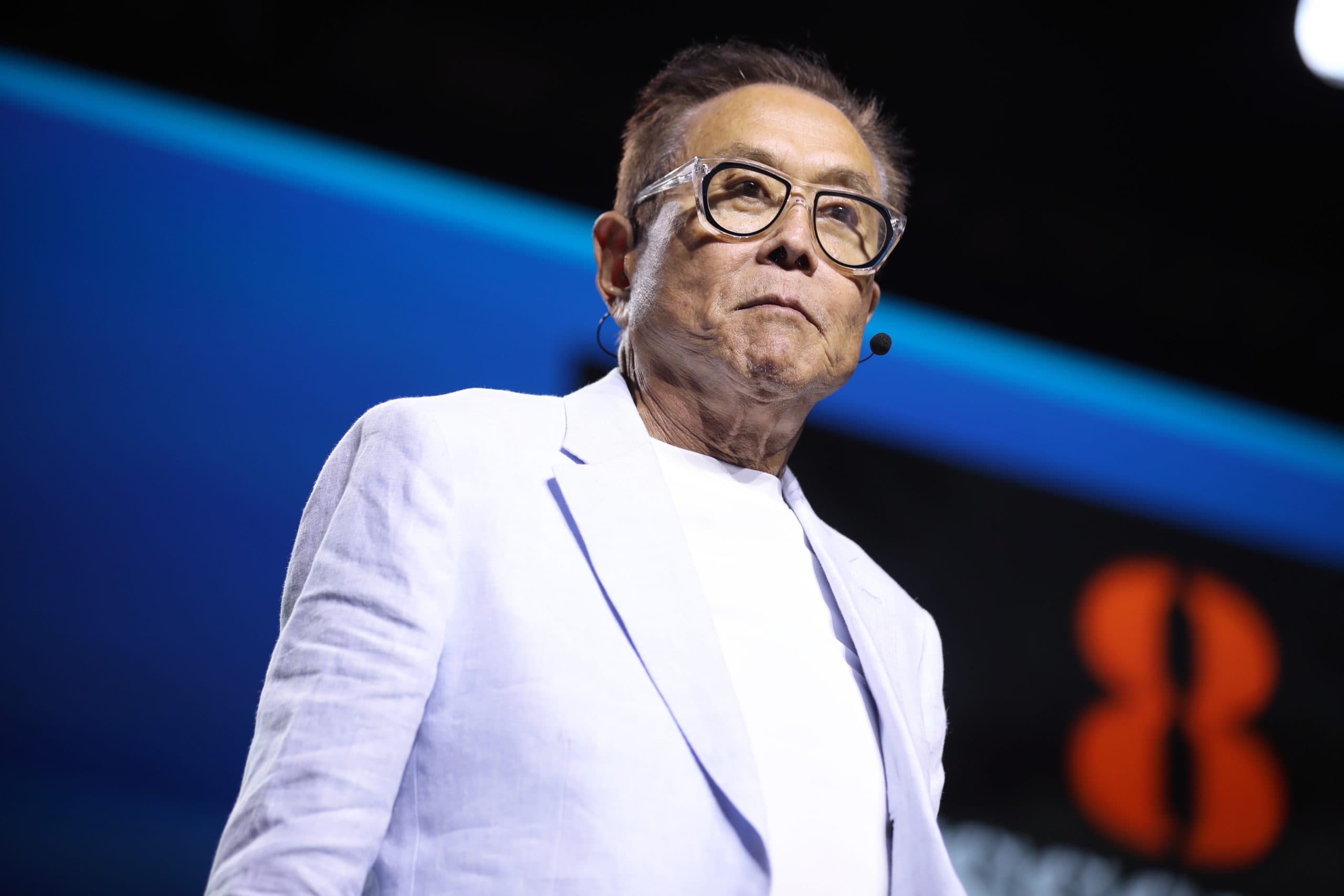 Eine Person (Robert Kiyosaki) mit Brille, weißem Hemd und leichter Jacke steht unter hellem Licht auf der Bühne und diskutiert leidenschaftlich über Bitcoin in ein Mikrofon-Headset.