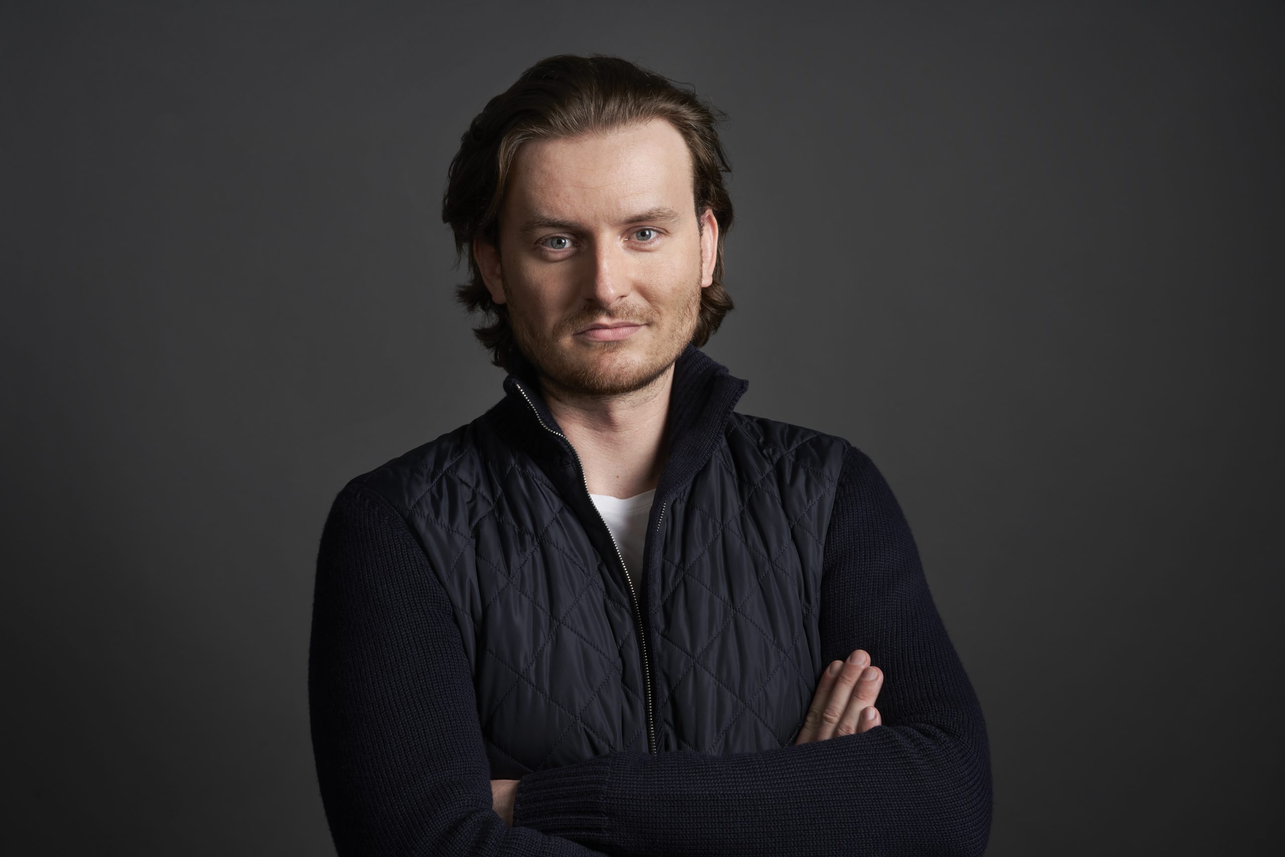 Eine Person (Eric Demuth) mit mittellangem Haar, die eine dunkle gesteppte Bitpanda-Jacke über einem Pullover trägt, steht mit verschränkten Armen vor einem dunkelgrauen Hintergrund.