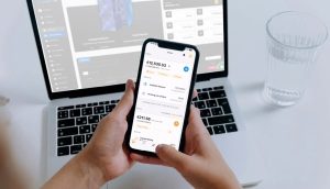 Eine Person hält ein Smartphone, auf dem die Finanz-App Millionminer mit Euro-Salden angezeigt wird, vor einem offenen Laptop und einem Glas Wasser auf einem weißen Schreibtisch.
