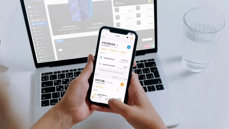 Eine Person hält ein Smartphone, auf dem die Finanz-App Millionminer mit Euro-Salden angezeigt wird, vor einem offenen Laptop und einem Glas Wasser auf einem weißen Schreibtisch.