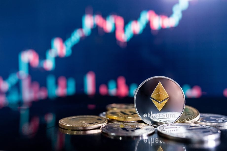 Mehrere Münzen, darunter eine Ethereum-Münze, liegen auf einer reflektierenden Oberfläche mit einem unscharfen Preisdiagramm der Kryptowährung im Hintergrund.