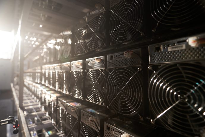 Reihen von Kryptowährungs-Mining-Rigs brummen unermüdlich in einem schwach beleuchteten Raum, ihre Kühlventilatoren surren leise. Etwas Licht fällt von links ein und wirft subtile Schatten auf die geordnete Ausrüstung, die für effiziente Mining-Operationen sorgt.
