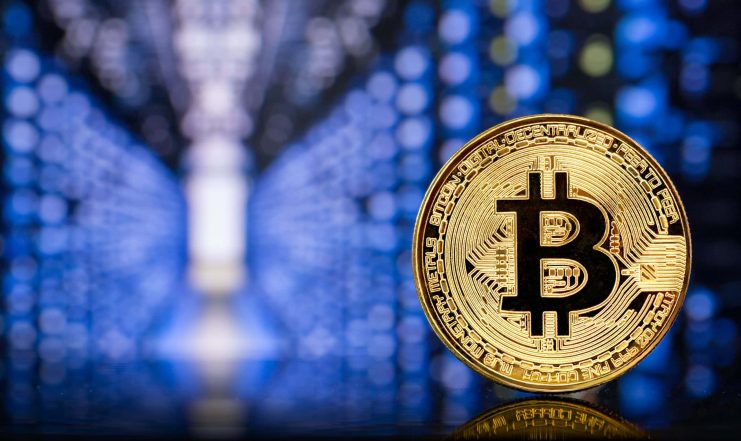 Eine physische Bitcoin-Münze steht aufrecht vor einem unscharfen Hintergrund aus blauen digitalen Datenmustern und symbolisiert die Verschmelzung von digitaler Währung und Technologie.
