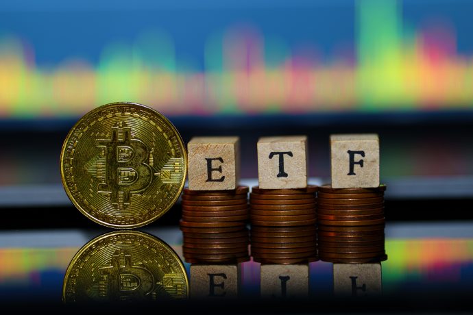 Eine physische Bitcoin-Münze steht neben Münzstapeln, die mit Holzklötzen mit der Aufschrift „ETF“ bedeckt sind, mit einem bunten Bitcoin-Handelsdiagramm im Hintergrund.