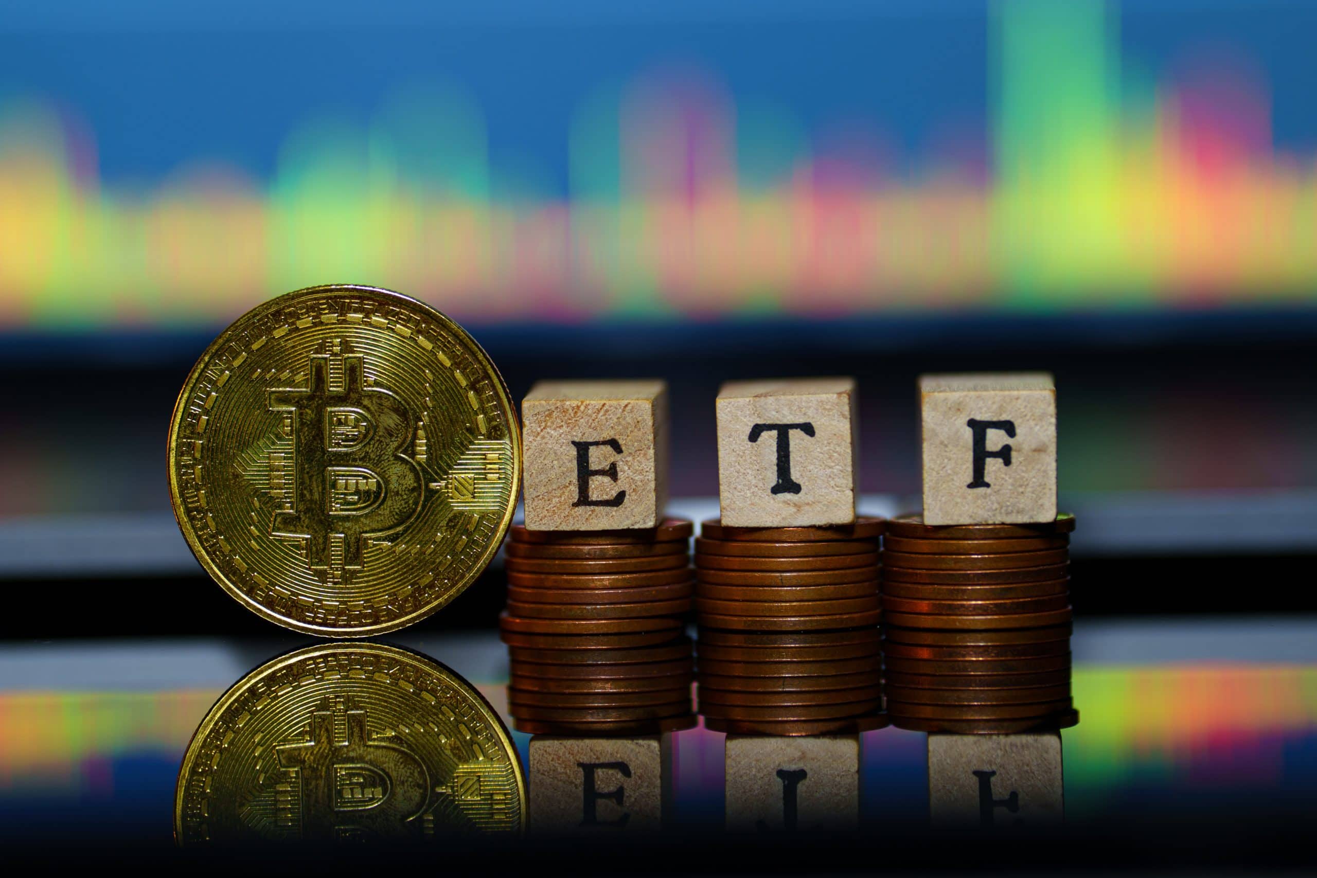 Eine physische Bitcoin-Münze steht neben Münzstapeln, die mit Holzklötzen mit der Aufschrift „ETF“ bedeckt sind, mit einem bunten Bitcoin-Handelsdiagramm im Hintergrund.