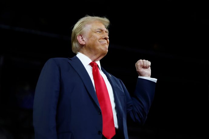 Der US-Präsident Donald Trump steht mit erhobener rechter Faust und einem leichten Lächeln vor einem dunklen Hintergrund.
