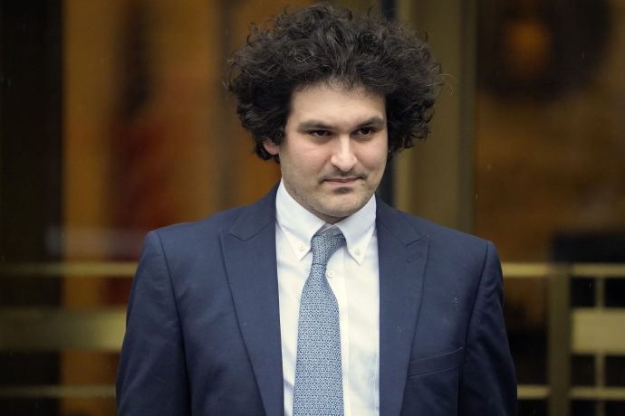 Eine Person mit lockigem Haar, die Sam Bankman-Fried ist, steht in einem blauen Anzug, einem weißen Hemd und einer gemusterten Krawatte vor einem leicht unscharfen Hintergrund.