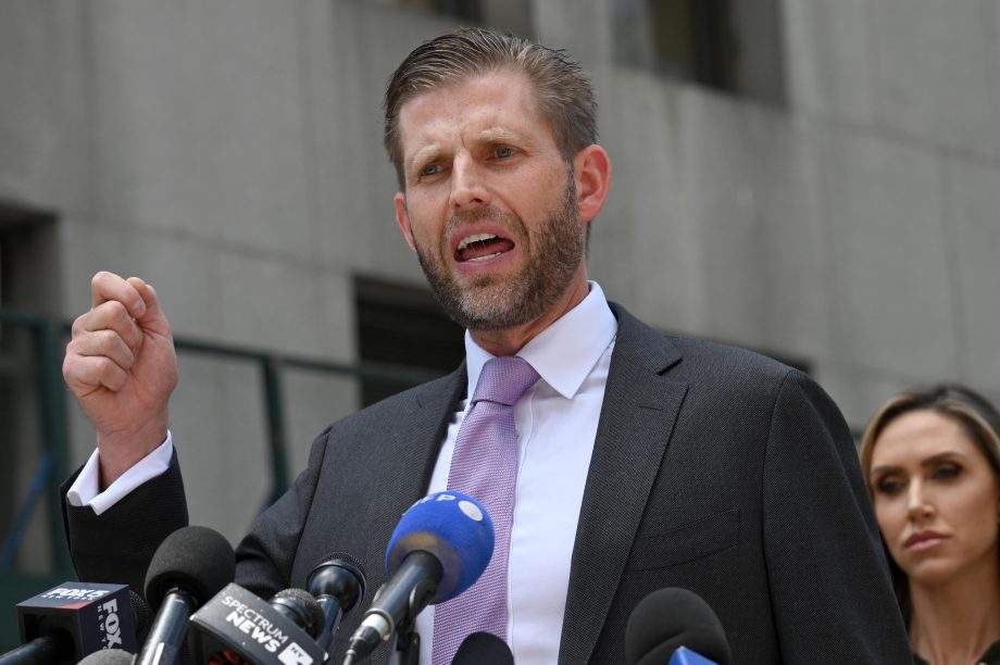 Ein Mann in Anzug und Krawatte spricht mit Nachdruck an einem Podium mit mehreren Mikrofonen, möglicherweise über Krypto-Themen im Freien in der Nähe eines Gebäudes, wobei eine weitere Person teilweise hinter ihm zu sehen ist.