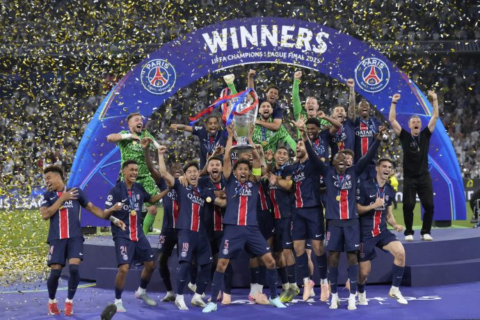 Die Spieler von Paris Saint-Germain feiern auf der Bühne mit der Trophäe unter fallendem Konfetti ihren Gewinn der UEFA Champions League, während über ihnen ein großes WINNERS-Schild und das Logo des Sponsors Krypto leuchten.