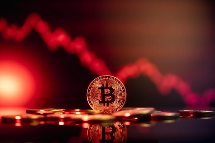 Eine Bitcoin-Münze liegt auf einer reflektierenden Oberfläche, im Hintergrund zeichnet sich ein rotes, nach unten gerichtetes Liniendiagramm ab, das einen kürzlichen Rückgang des Wertes der Kryptowährung signalisiert.