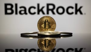 Eine Bitcoin-Münze steht aufrecht vor dem verschwommenen BlackRock-Logo, zwei weitere Bitcoin-Münzen liegen flach auf einer reflektierenden Oberfläche.