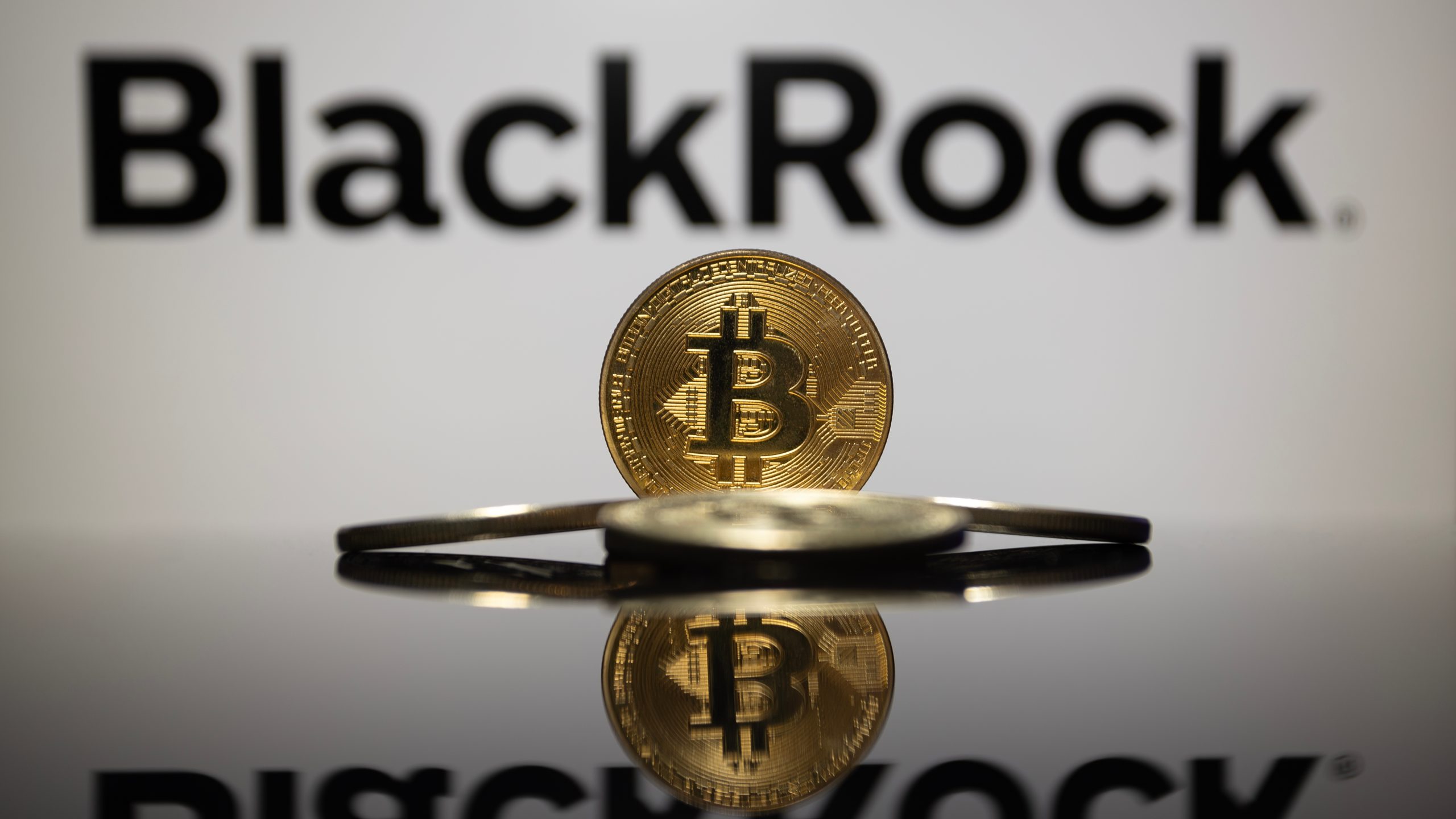 Eine Bitcoin-Münze steht aufrecht vor dem verschwommenen BlackRock-Logo, zwei weitere Bitcoin-Münzen liegen flach auf einer reflektierenden Oberfläche.