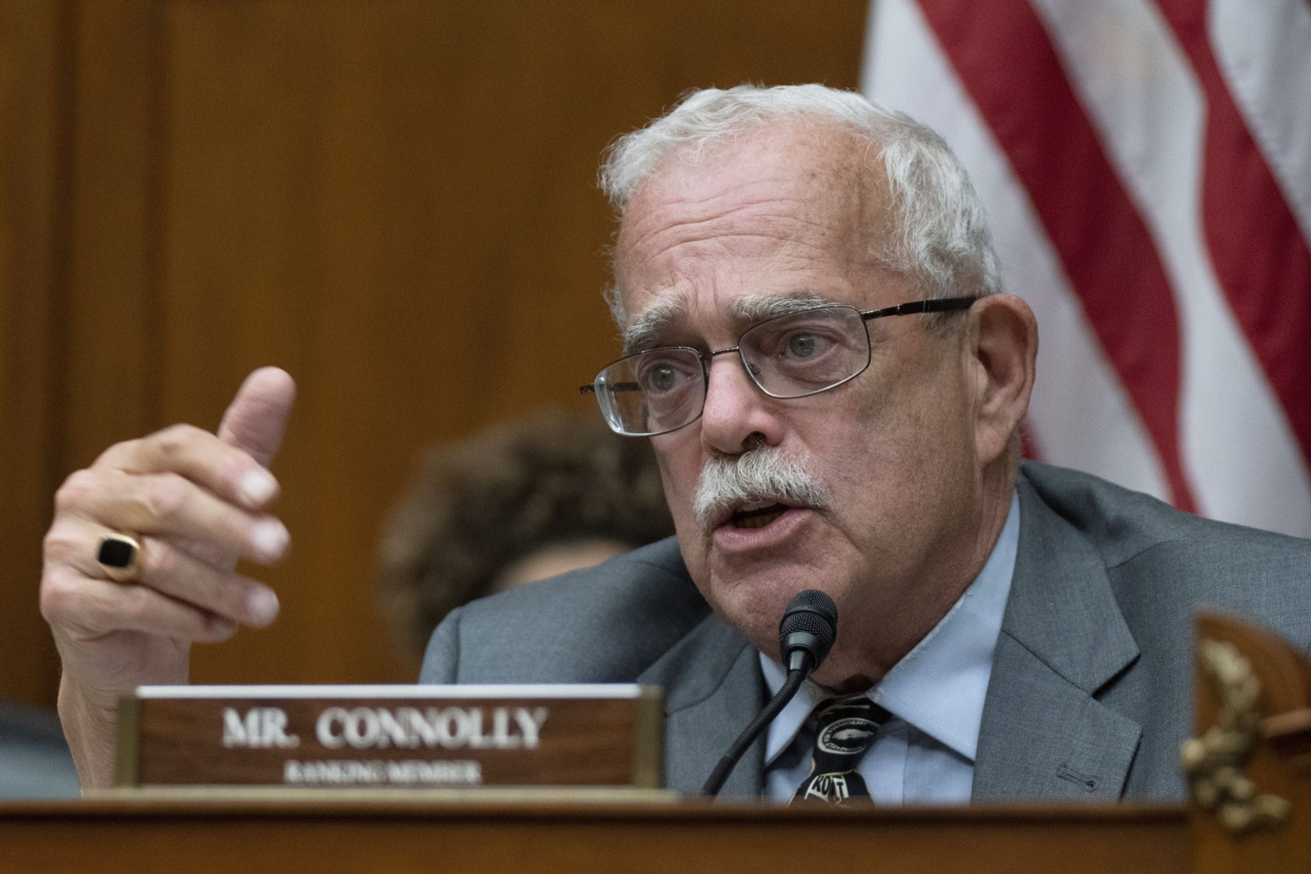Gerald Connolly spricht bei einem Meeting in ein Mikrofon. Er sitzt hinter einem Namensschild mit der Aufschrift „Mr. Connolly“, im Hintergrund ist die US-Flagge zu sehen.