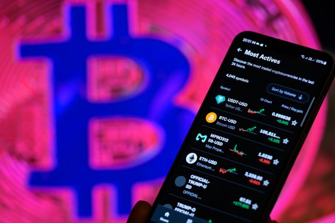 Auf einem Smartphone werden Handelsdaten zu Kryptowährungen angezeigt. Im Hintergrund ist ein buntes, verschwommenes Bitcoin-Logo zu sehen – ein unverzichtbares Tool für die Verwaltung von Bitcoin-Portfolios, selbst bei einem Minus von 23 Prozent.