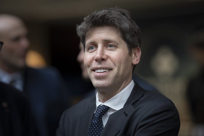 Sam Altman steht selbstbewusst in einem schwach beleuchteten Innenbereich.