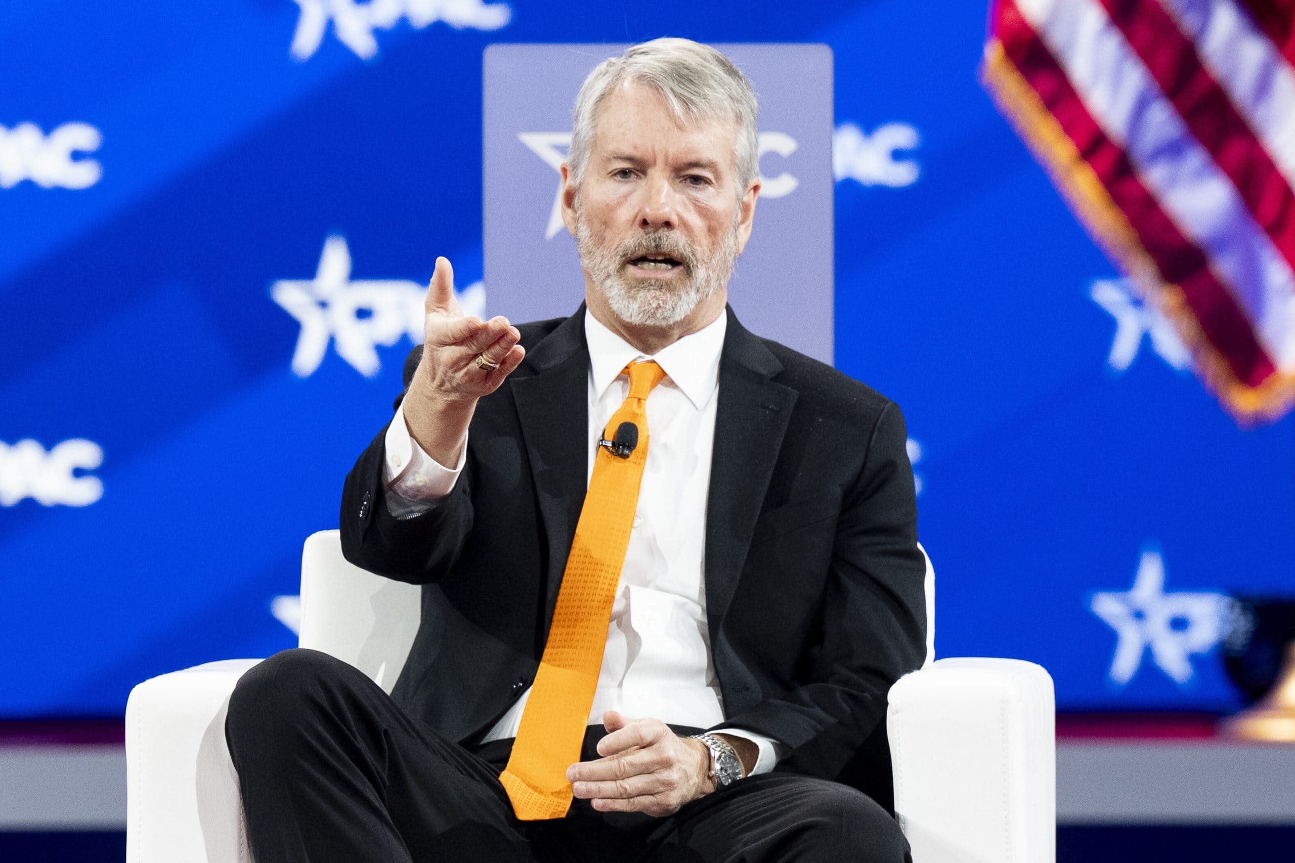 Ein Mann in Anzug und orangefarbener Krawatte, der Michael Saylor ähnelt, gestikuliert, während er auf der Bühne einer Konferenz spricht. Im Hintergrund ist teilweise eine amerikanische Flagge zu sehen.