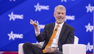 Ein Mann in Anzug und orangefarbener Krawatte sitzt auf einem Bühnenstuhl und gestikuliert mit der Hand. Auf dem blauen Hintergrund ist ein weißes Logo zu sehen, das subtil auf den Einfluss von Bitcoin hinweist.