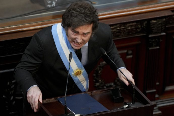 Javier Milei mit einer zeremoniellen Schärpe spricht leidenschaftlich an einem Holzpult in einem formellen Rahmen.