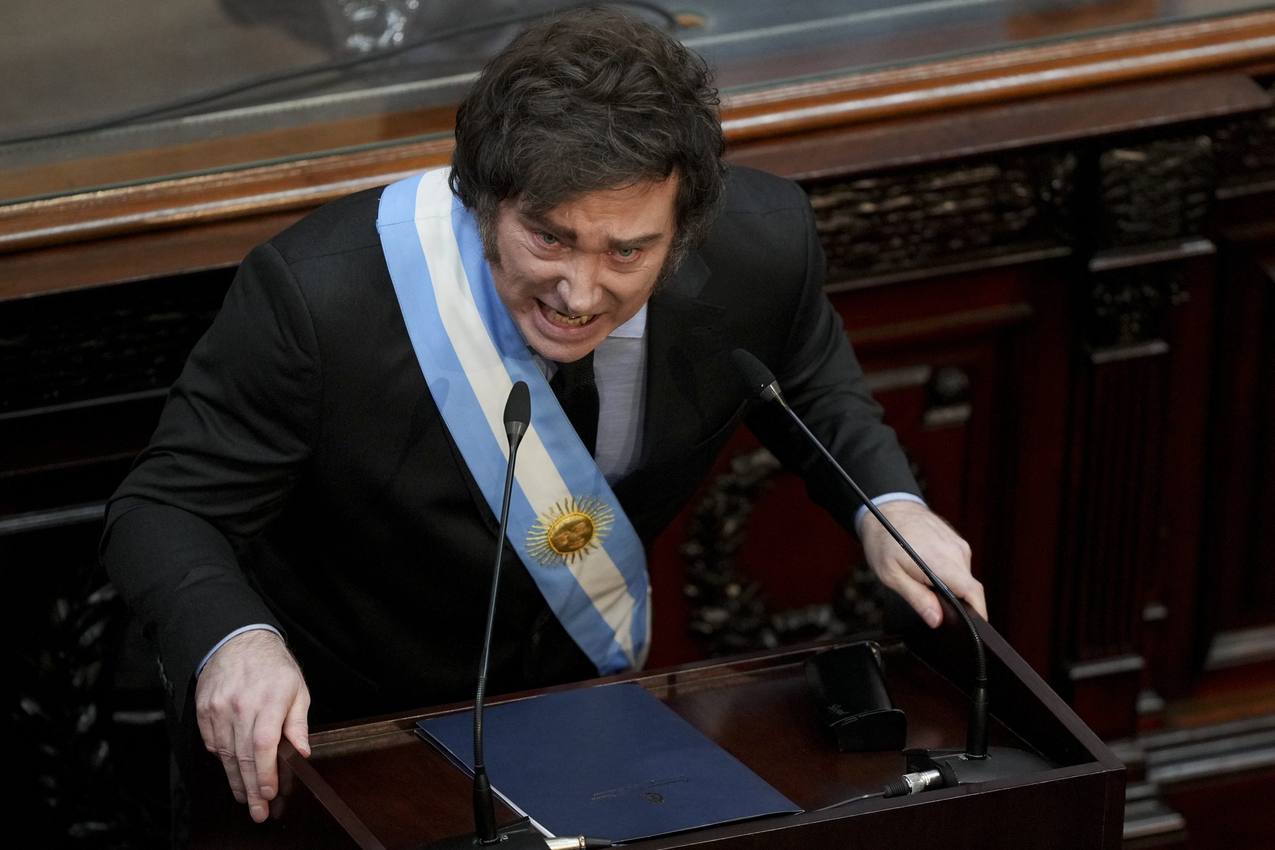 Javier Milei mit einer zeremoniellen Schärpe spricht leidenschaftlich an einem Holzpult in einem formellen Rahmen.