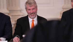 Michael Saylor sitzt lächelnd an einem Tisch. Neben ihm sind zwei weitere Personen teilweise zu sehen
