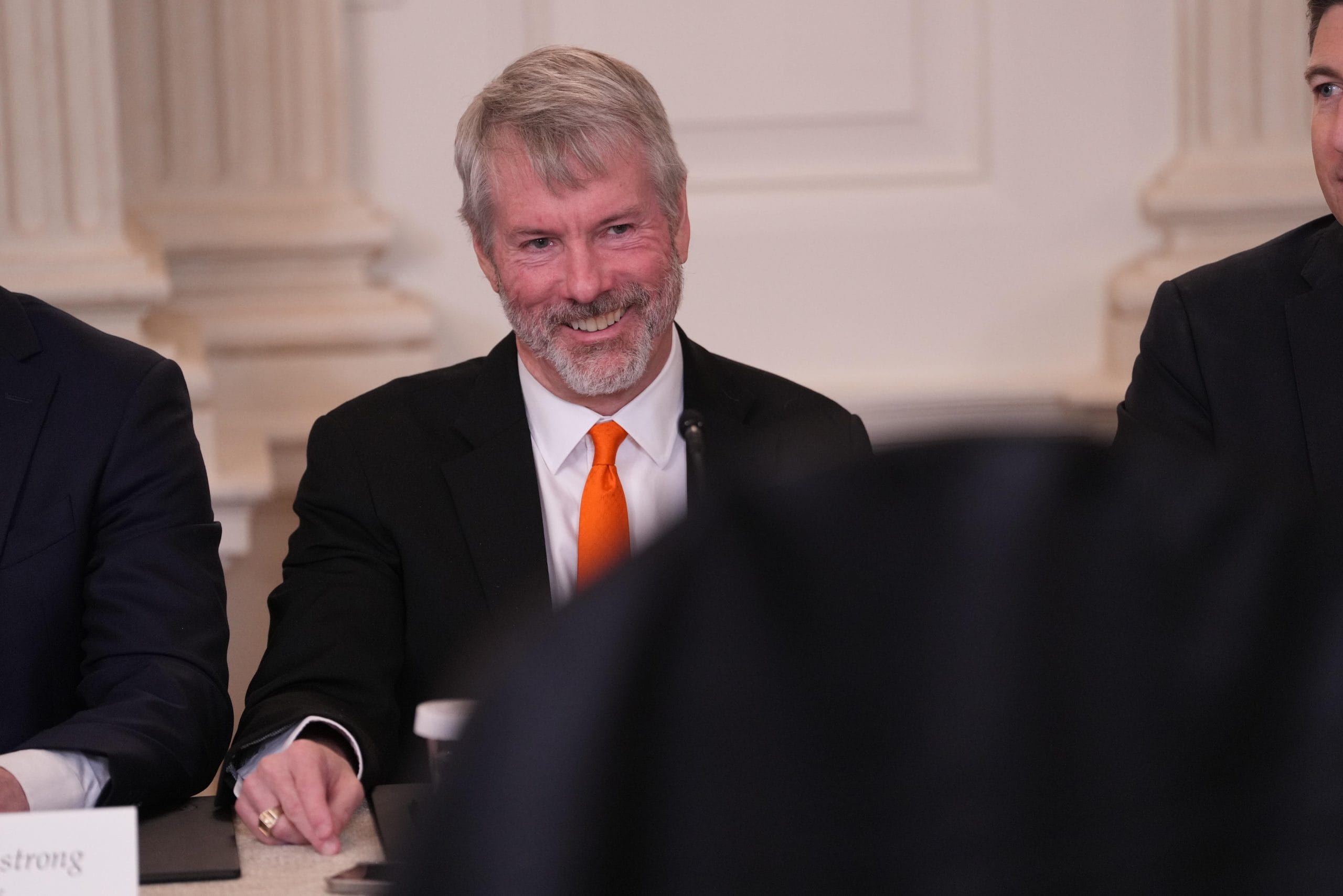 Michael Saylor sitzt lächelnd an einem Tisch. Neben ihm sind zwei weitere Personen teilweise zu sehen