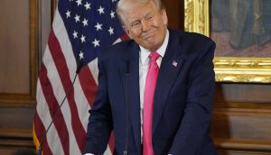 Donald Trump in Anzug und rosa Krawatte steht an einem Podium mit Mikrofonen, was an Trumps Stil erinnert, und spricht vor einer amerikanischen Flagge und einem gerahmten Gemälde zu einem Publikum.