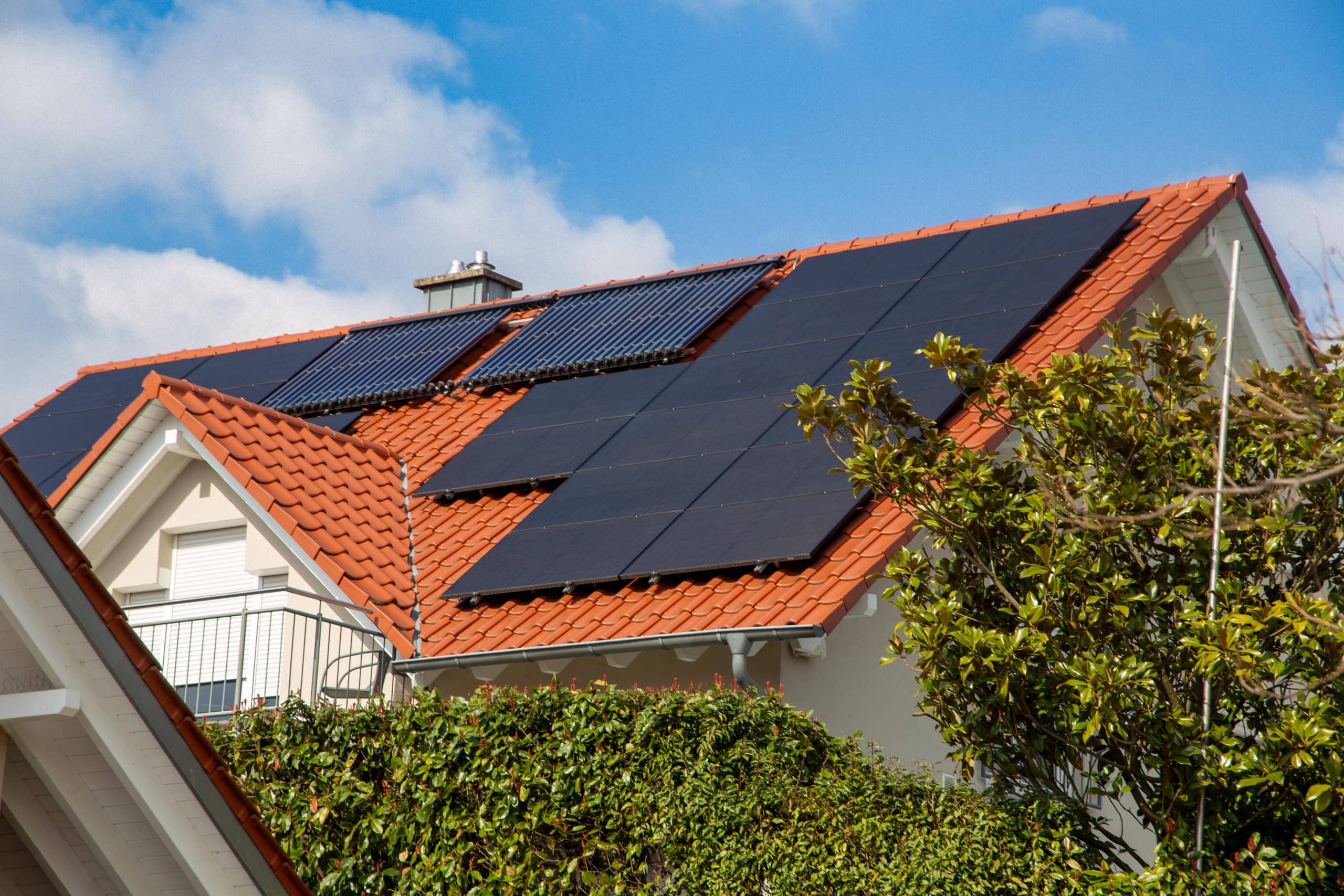 Ein Haus mit einem roten Ziegeldach, das für den Selbstversorger-Lebensstil konzipiert wurde, ist mit mehreren Solarmodulen ausgestattet und von üppigem Grün umgeben, vor einem teilweise bewölkten Himmel.