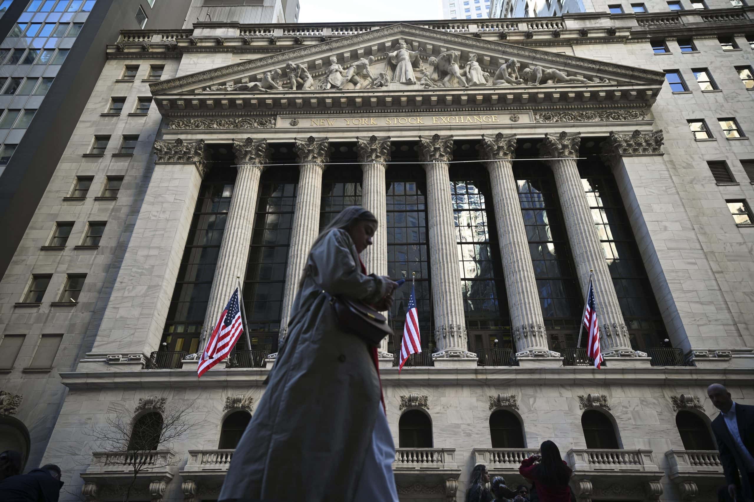 Person, die vor dem Gebäude der New Yorker Börse geht, das eine klassische Fassade mit Säulen und US-Flaggen aufweist.
