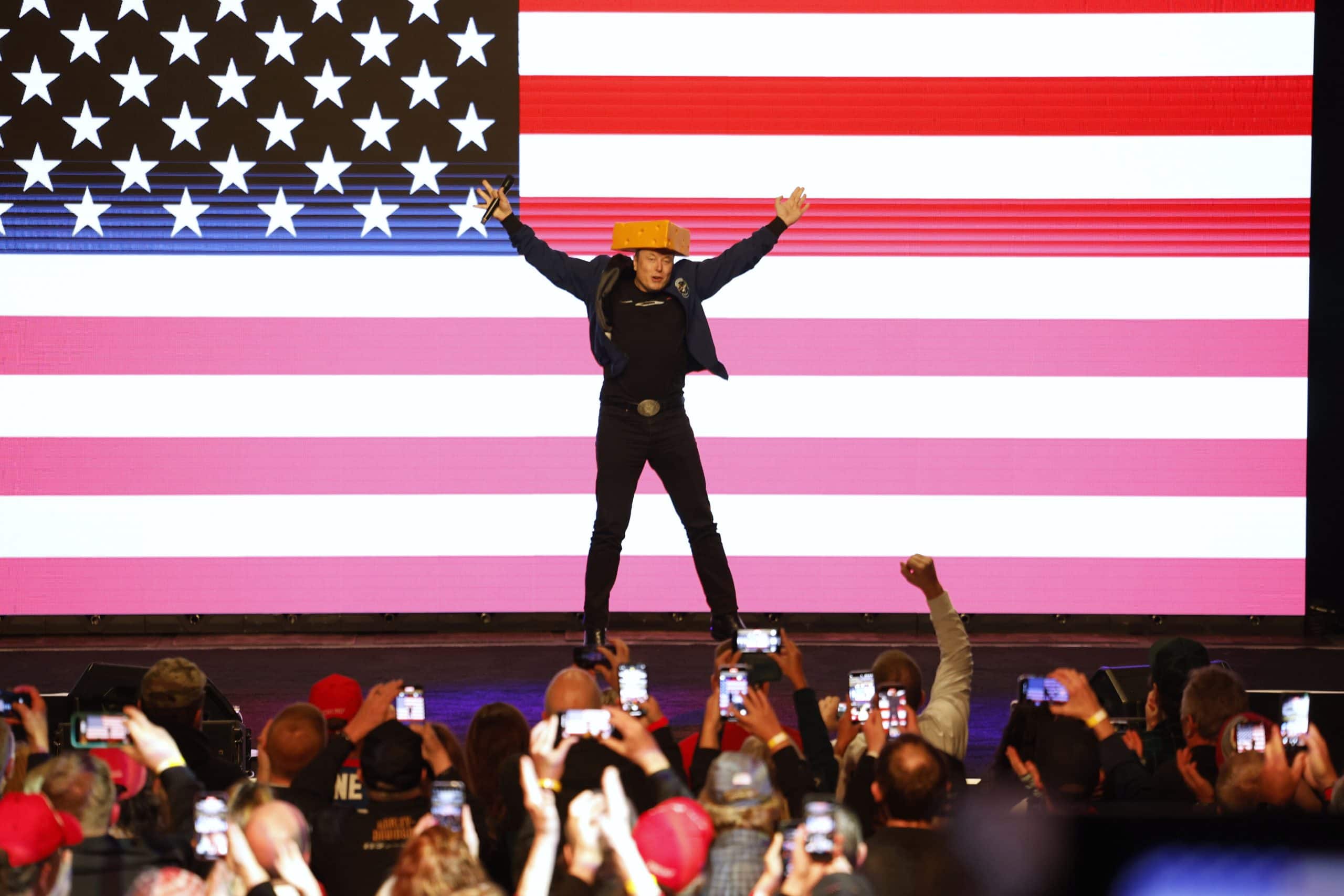 Elon Musk auf der Bühne trägt einen Käsehut und erhebt die Arme vor einer großen Ausstellung amerikanischer Flaggen. Zuschauer machen Fotos mit ihren Handys.