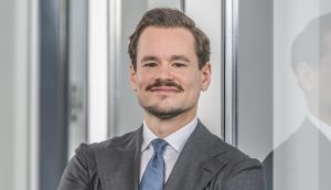 Ein Geschäftsführer in Anzug und Krawatte steht selbstbewusst mit verschränkten Armen und einem leichten Lächeln vor einer spiegelnden Glasfläche im Innenbereich.
