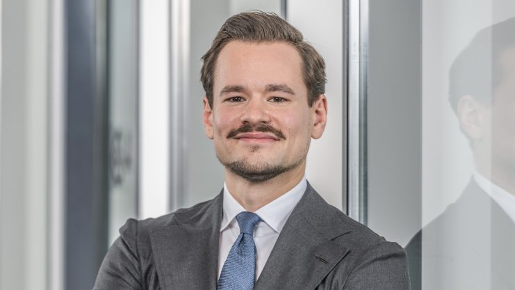 Ein Geschäftsführer in Anzug und Krawatte steht selbstbewusst mit verschränkten Armen und einem leichten Lächeln vor einer spiegelnden Glasfläche im Innenbereich.