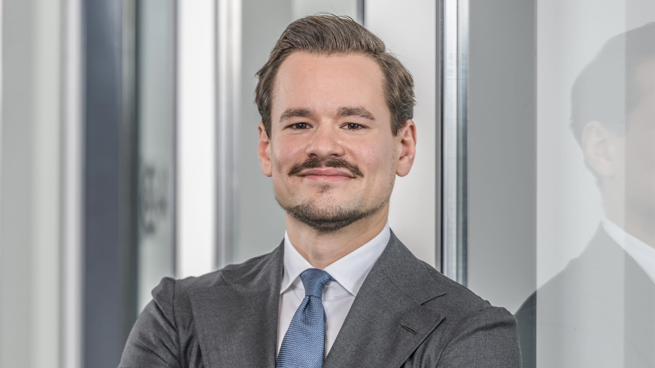 Ein Geschäftsführer in Anzug und Krawatte steht selbstbewusst mit verschränkten Armen und einem leichten Lächeln vor einer spiegelnden Glasfläche im Innenbereich.