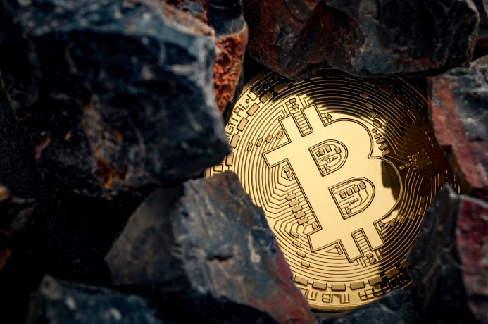 Eine goldfarbene Bitcoin-Münze ist teilweise von dunklen Felsen umgeben, was den Reiz von Bitcoin inmitten des zerklüfteten Geländes unterstreicht.