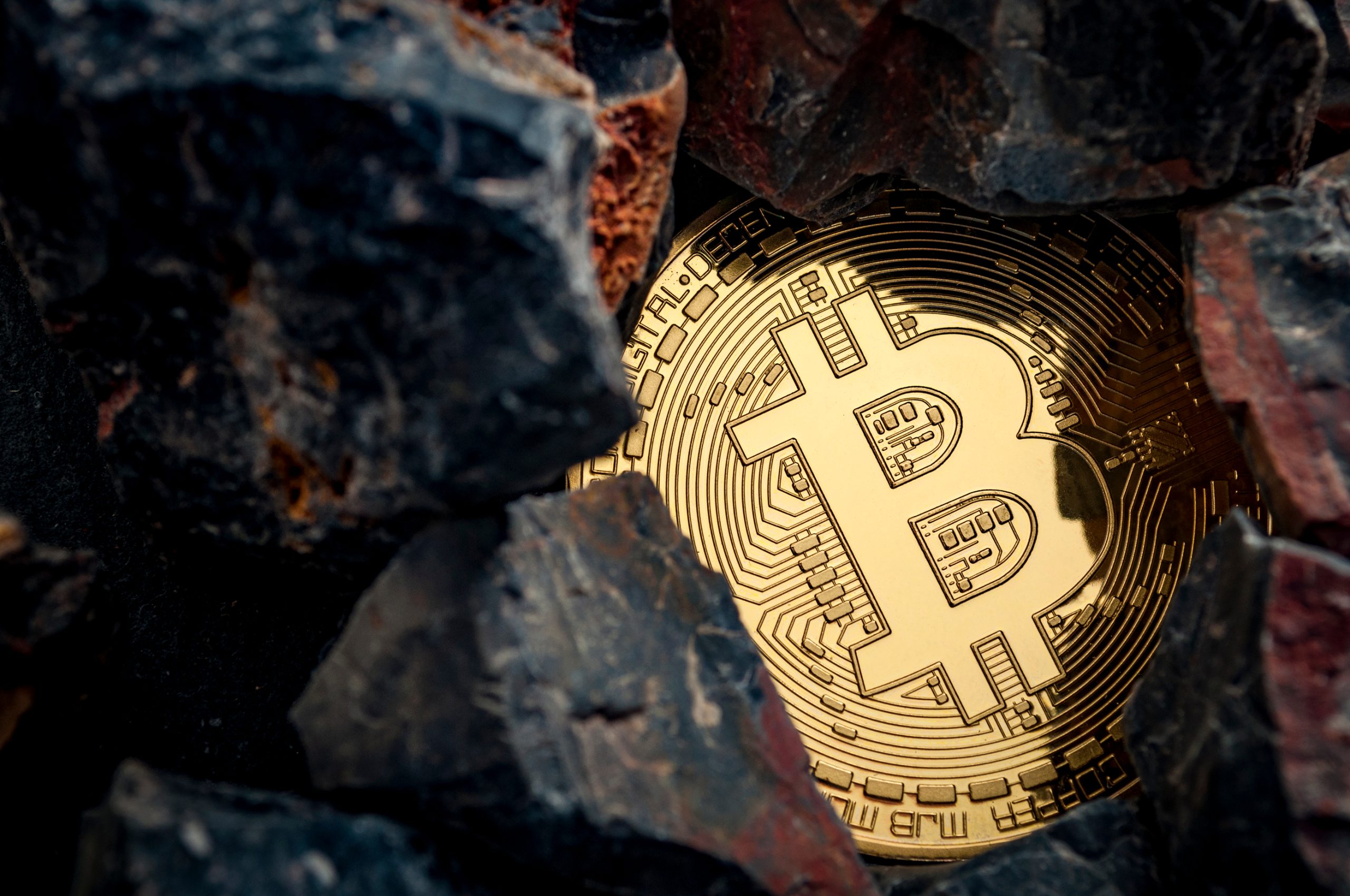 Eine goldfarbene Bitcoin-Münze ist teilweise von dunklen Felsen umgeben, was den Reiz von Bitcoin inmitten des zerklüfteten Geländes unterstreicht.