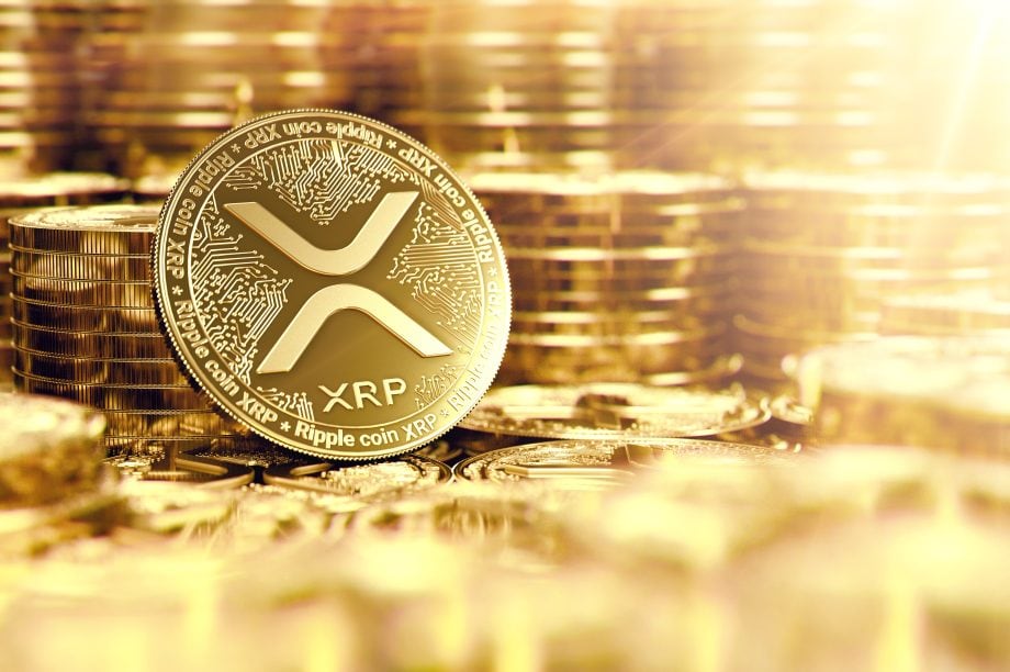 Eine goldfarbene physische Münze mit dem XRP-Logo steht aufrecht zwischen Stapeln ähnlicher Münzen und wird im Hintergrund von einem hellen, goldenen Licht beleuchtet.