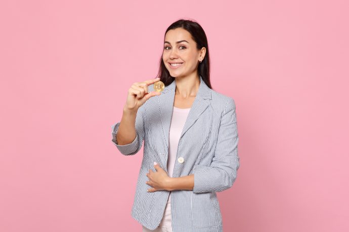 Eine Frau im gestreiften Blazer hält stolz eine Bitcoin-Münze in der Hand. Ihr Lächeln strahlt vor dem rosa Hintergrund und verkörpert den Geist von „Frauen investieren“.