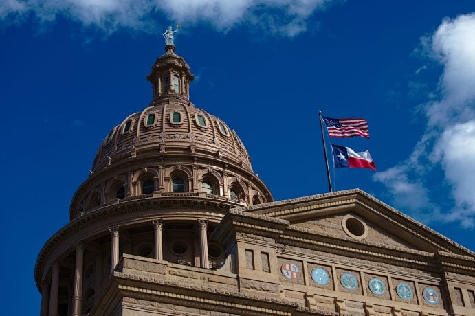 Die Kuppel des texanischen Kapitols ragt stolz vor einem blauen Himmel empor, daneben wehen die Flaggen der USA und von Texas, während der Texanische Senat über einen neuen Gesetzentwurf nachdenkt.