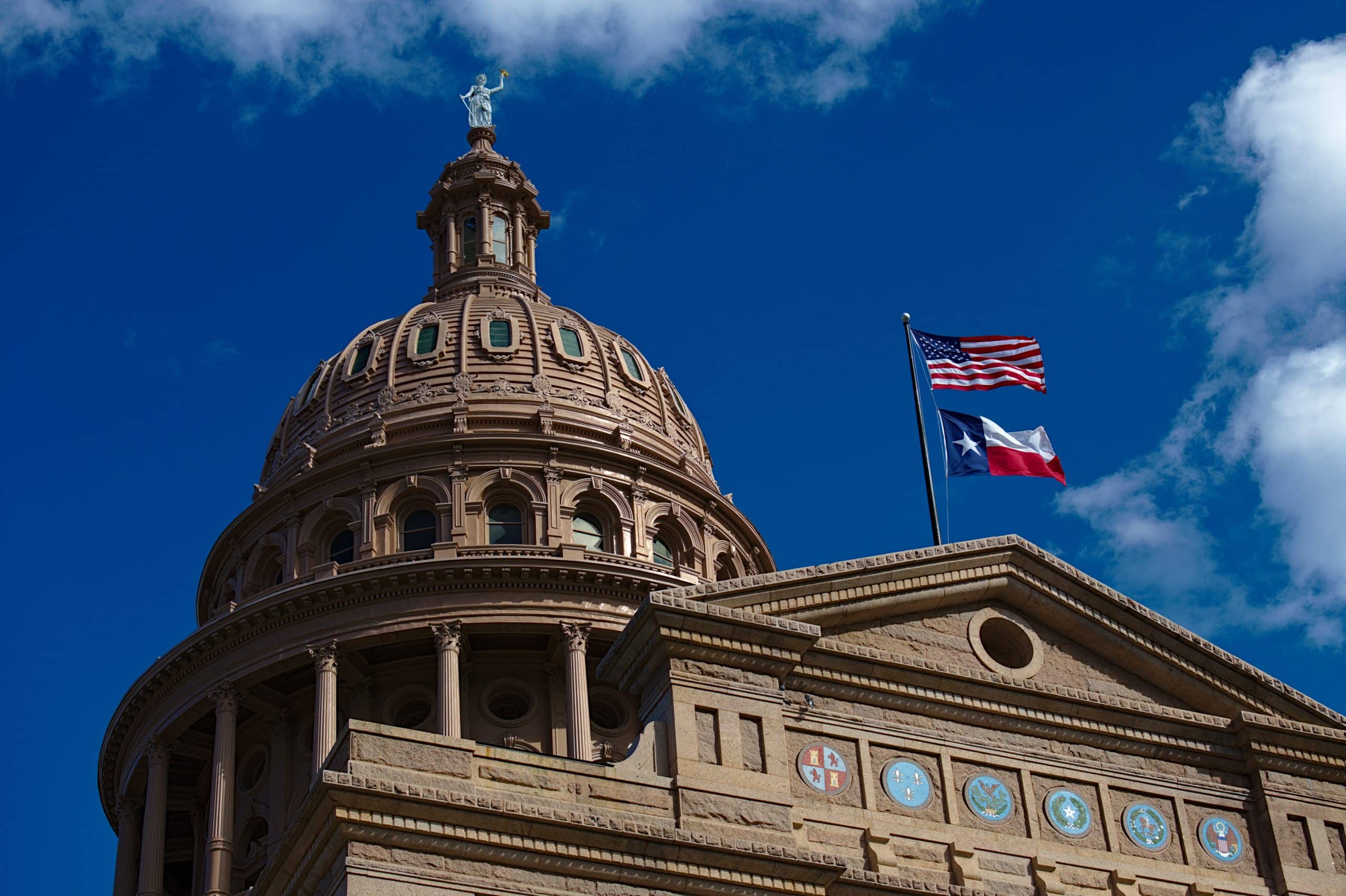 Die Kuppel des texanischen Kapitols ragt stolz vor einem blauen Himmel empor, daneben wehen die Flaggen der USA und von Texas, während der Texanische Senat über einen neuen Gesetzentwurf nachdenkt.