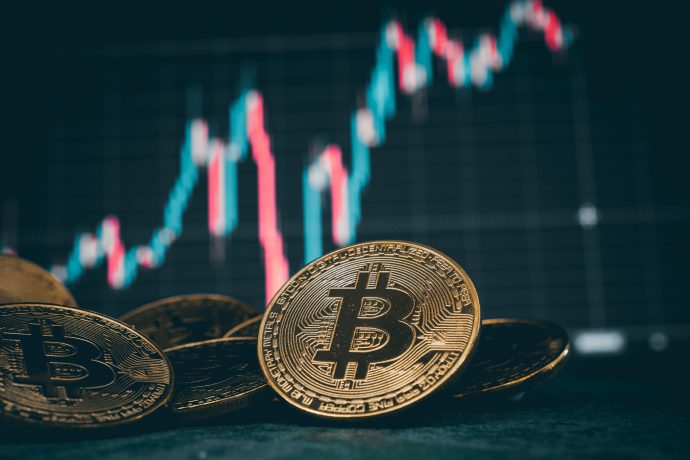 Im Vordergrund werden goldene Bitcoin-Münzen angezeigt, im Hintergrund ein unscharfes Finanzaktiendiagramm.